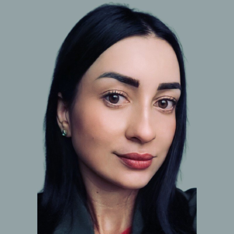 Foto de Débora Prudente, com cabelos longos pretos e jaqueta preta, destacando-se em um fundo cinza