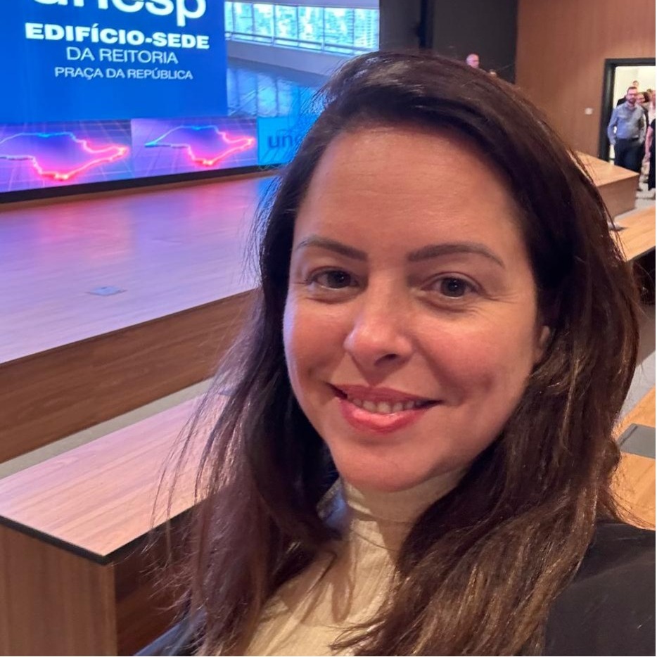 Foto de Luciana, com cabelos longos castanhos, sorrindo em frente a uma tela grande com a frase unesp